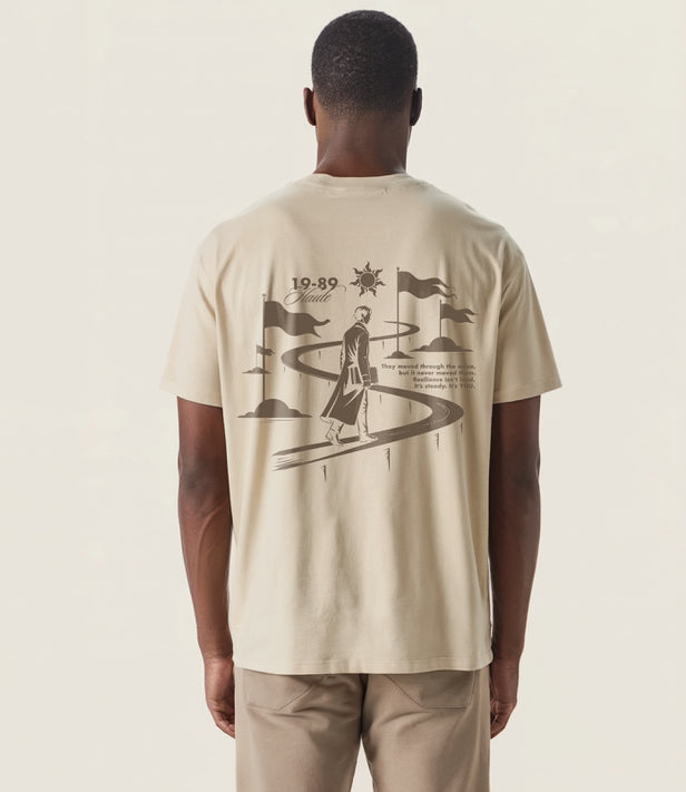 Back view of sandstone oversized T-shirt featuring 19-89 Haute graphic design of a man walking along a winding path with flags and sun symbol, representing resilience and journey.