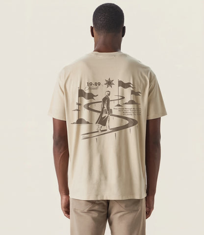 Back view of sandstone oversized T-shirt featuring 19-89 Haute graphic design of a man walking along a winding path with flags and sun symbol, representing resilience and journey.